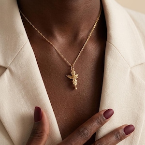 Puede incluir: Un collar dorado con un colgante de querub&iacute;n alado. El querub&iacute;n est&aacute; de pie, con detalles finos. La cadena es delicada. El fondo es una blazer color crema.