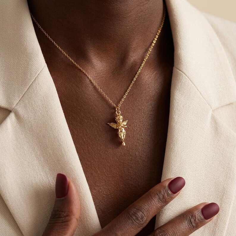 Puede incluir: Un collar dorado con un colgante de querub&iacute;n alado. El querub&iacute;n est&aacute; de pie, con detalles finos. La cadena es delicada. El fondo es una blazer color crema.