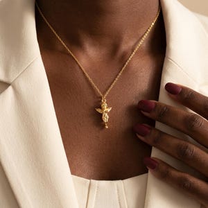 Puede incluir: Un collar dorado con un colgante de querub&iacute;n. El colgante representa un peque&ntilde;o &aacute;ngel con alas, suspendido de una fina cadena. El collar se lleva sobre una blazer color crema. Las u&ntilde;as de la persona est&aacute;n pintadas de color rojo oscuro.