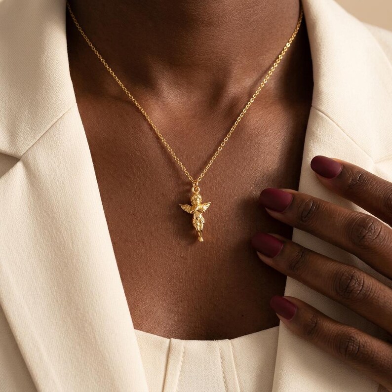 Puede incluir: Un collar dorado con un colgante de querub&iacute;n. El colgante representa un peque&ntilde;o &aacute;ngel con alas, suspendido de una fina cadena. El collar se lleva sobre una blazer color crema. Las u&ntilde;as de la persona est&aacute;n pintadas de color rojo oscuro.