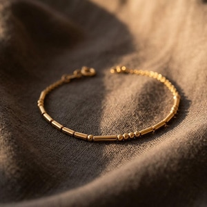 Könnte beinhalten: Ein goldfarbenes Armband mit einer zarten Kette, die aus abwechselnden kleinen zylindrischen Stäbchen und runden Perlen besteht. Das Armband verfügt über einen runden Verschluss an einem Ende und weist ein poliertes Metallfinish auf.