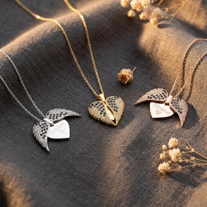 Pode incluir: Três colares com pingentes em forma de coração com asas de anjo. Um é prateado com pedras pretas e brancas, outro é dourado com pedras pretas e brancas, e o terceiro é em ouro rosa com pedras pretas e brancas. Cada colar tem um nome gravado no coração.