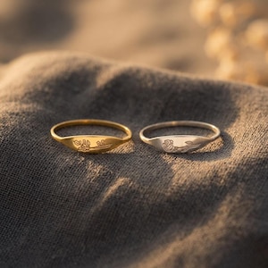 Könnte beinhalten: Zwei dünne Metallringe, einer goldfarben und einer silberfarben, mit jeweils einer flachen, ovalen Oberfläche, die mit einem zarten Blumenmuster graviert ist. Der goldene Ring zeigt einen Stängel mit Blättern, der silberne eine einzelne Rose.