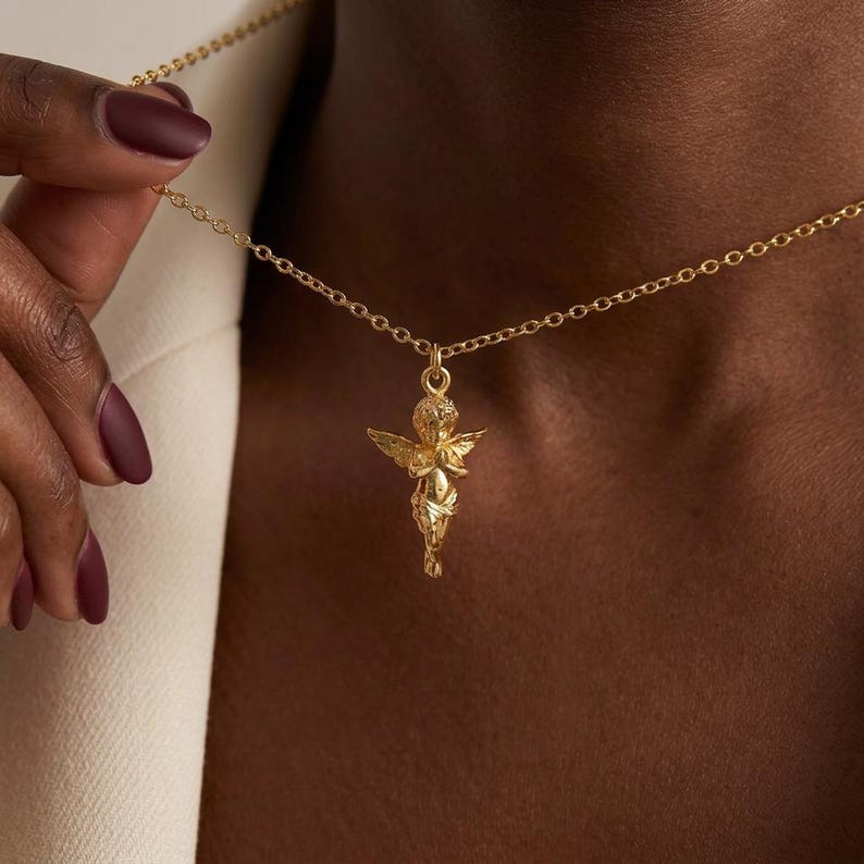Puede incluir: Un collar dorado con un colgante de querub&iacute;n. El querub&iacute;n tiene alas detalladas y est&aacute; suspendido de una fina cadena. El fondo es marr&oacute;n oscuro, lo que acent&uacute;a el oro del collar.