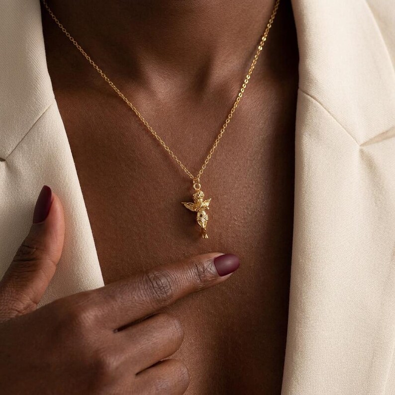 Puede incluir: Un collar dorado con un peque&ntilde;o colgante de querub&iacute;n. El querub&iacute;n tiene alas extendidas y est&aacute; detallado con peque&ntilde;os adornos. La cadena del collar es delicada y tambi&eacute;n dorada. El fondo muestra una blazer color crema.