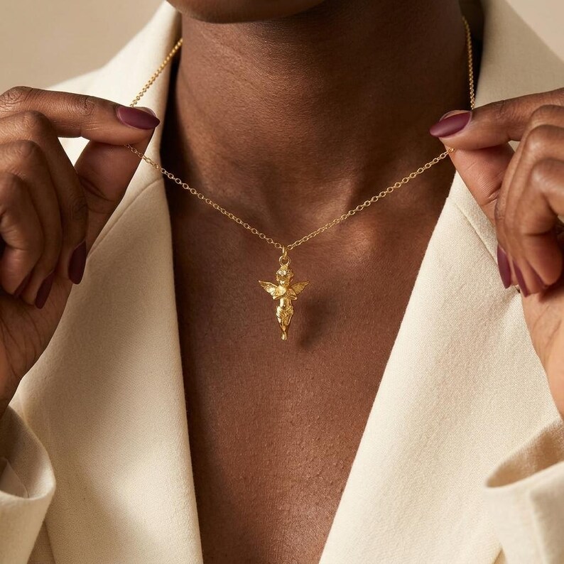 Puede incluir: Un collar dorado con un colgante que representa un peque&ntilde;o &aacute;ngel con las alas extendidas. El &aacute;ngel cuelga de una fina cadena. El collar se lleva sobre una blazer color crema.