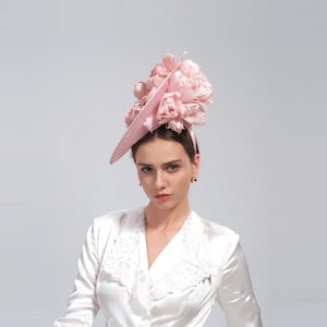 May include: A light pink fascinator with a wide brim and a large floral arrangement. The headpiece is secured with a thin headband. The woman in the image is wearing a white satin blouse with lace trim.