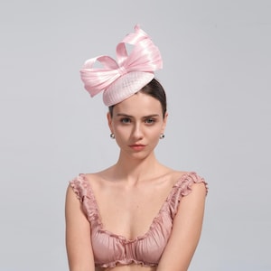 Puede incluir: Un sombrero rosa claro con un gran lazo en la parte superior. El sombrero es de color rosa pálido y tiene un aspecto texturizado. El lazo está hecho de un material brillante, similar al satén. El sombrero se lleva en la cabeza.