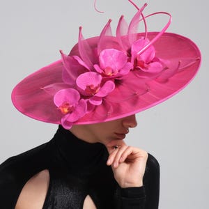 Könnte beinhalten: Ein leuchtend pinkfarbener Hut mit breiter Krempe, verziert mit großen, leuchtend rosa Orchideenblüten und zarten, transparenten Akzenten. Der Hut ist ein Statement-Stück, ideal für besondere Anlässe.