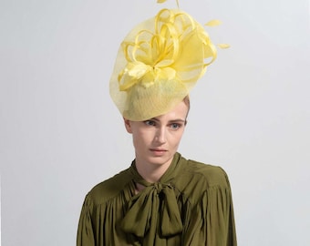 May include: A bright yellow fascinator with a floral design. The headpiece features a mesh veil and is adorned with fabric petals. The woman in the image is wearing an olive green blouse.