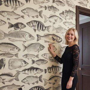 Könnte beinhalten: Ein Badezimmer mit Fischmuster-Tapete. Die Tapete zeigt schwarz-weiße Illustrationen verschiedener Fischarten auf cremefarbenem Hintergrund. Eine Frau in einem schwarzen Kleid steht lächelnd an der Wand. Eine gestreifte Bank und eine dunkelbraune Tür sind ebenfalls zu sehen.