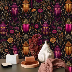 May include: A dark background with a repeating pattern of gold and magenta beetles and floral elements. The foreground features a white vase, a pink towel, soap, and a small black box.