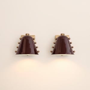 Op de afbeelding: Twee bijpassende wandkandelaars met een donkerbruine, klokvormige kap. Elke wandkandelaar heeft een messingkleurige montageplaat en decoratieve messingkleurige bollen. De wandkandelaars zijn op een lichtgekleurde muur gemonteerd.