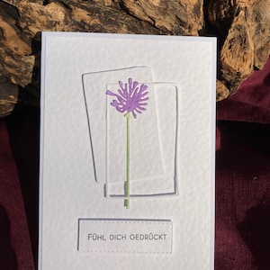 May include: A white greeting card with a textured surface. It features a purple flower design within two clear, rectangular frames. The text "F&Uuml;HL DICH GEDR&Uuml;CKT" is displayed at the bottom. The background includes wood and a burgundy fabric.