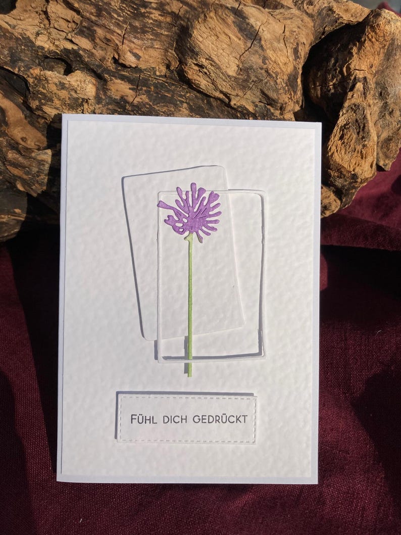 May include: A white greeting card with a textured surface. It features a purple flower design within two clear, rectangular frames. The text "F&Uuml;HL DICH GEDR&Uuml;CKT" is displayed at the bottom. The background includes wood and a burgundy fabric.