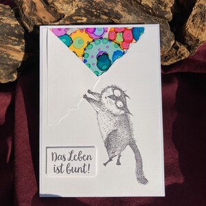 May include: A white greeting card with a whimsical illustration of a cat holding onto a colorful balloon. The card features the German phrase "Das Leben ist bunt!" which translates to "Life is colorful!"