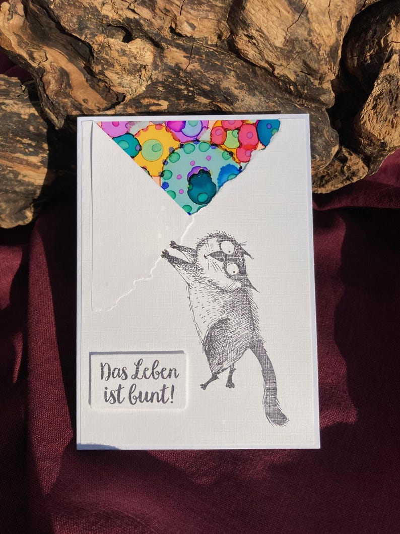 May include: A white greeting card with a whimsical illustration of a cat holding onto a colorful balloon. The card features the German phrase "Das Leben ist bunt!" which translates to "Life is colorful!"