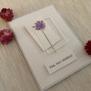 May include: A handmade greeting card with a purple flower design and the text "F&uuml;hl dich gedr&uuml;ckt". The card is decorated with three dried red flowers. The card is on a textured white surface.