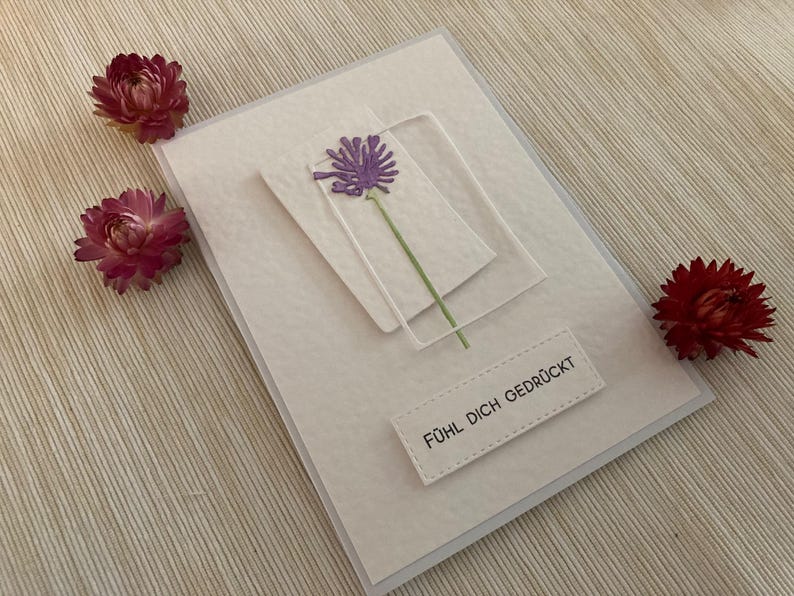 May include: A handmade greeting card with a purple flower design and the text "F&uuml;hl dich gedr&uuml;ckt". The card is decorated with three dried red flowers. The card is on a textured white surface.