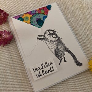 May include: A greeting card with a whimsical cat illustration, the text "Das Leben ist bunt!" is printed on a white rectangle. The envelope has a colorful abstract design. Pink and yellow flowers are placed around the card.