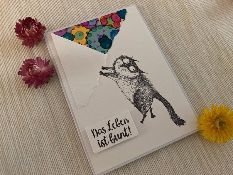 May include: A greeting card with a whimsical cat illustration, the text "Das Leben ist bunt!" is printed on a white rectangle. The envelope has a colorful abstract design. Pink and yellow flowers are placed around the card.