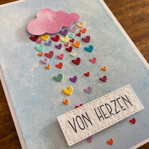 Könnte beinhalten: Eine handgefertigte Grußkarte mit hellblauem Hintergrund und einer rosa Wolke. Bunte herzförmige Verzierungen fallen aus der Wolke. Der Text "VON HERZEN" ist auf einem weißen rechteckigen Aufnäher abgebildet.