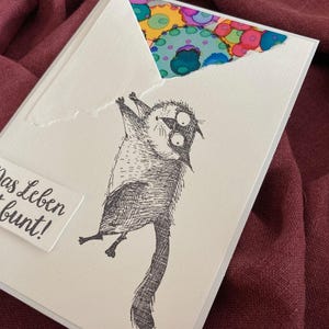 May include: A white greeting card with a whimsical illustration of a cat hanging upside down. The card features the German phrase "Das Leben ist bunt!" and a colorful patterned design peeking from the torn top.