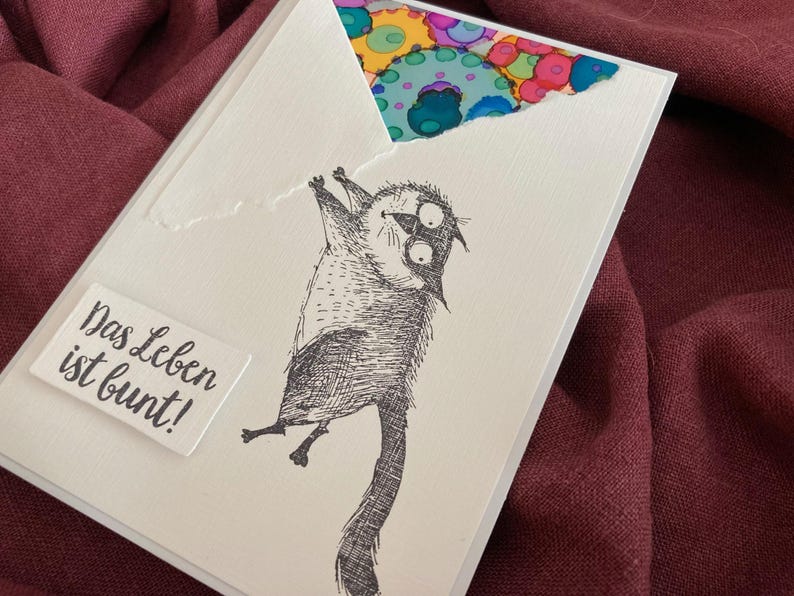 May include: A white greeting card with a whimsical illustration of a cat hanging upside down. The card features the German phrase "Das Leben ist bunt!" and a colorful patterned design peeking from the torn top.