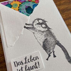 May include: A white greeting card with a whimsical black and white illustration of a cat-like creature reaching for a colorful design peeking out from behind a torn envelope. The text "Das Leben ist bunt!" is on a small white rectangle.