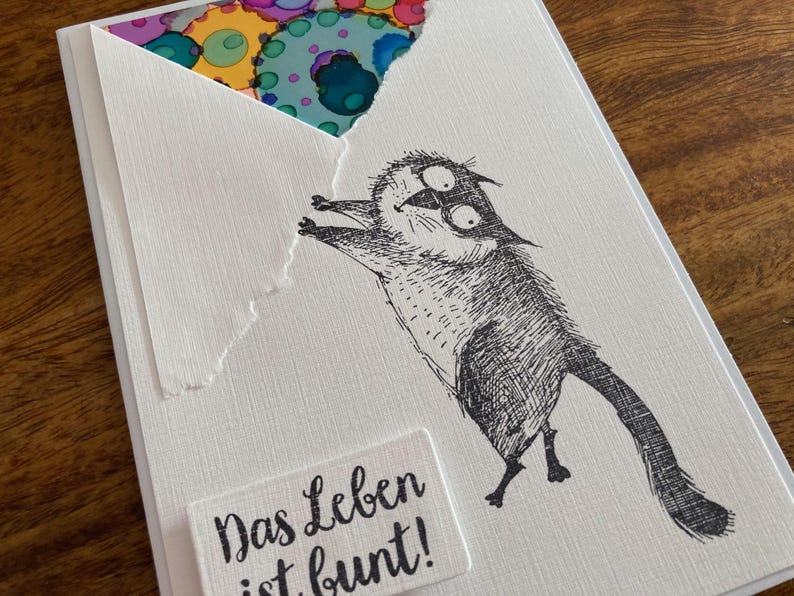 May include: A white greeting card with a whimsical black and white illustration of a cat-like creature reaching for a colorful design peeking out from behind a torn envelope. The text "Das Leben ist bunt!" is on a small white rectangle.