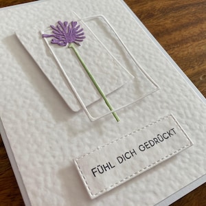 May include: A white greeting card with a textured surface, featuring a purple flower with a green stem and the German text "F&Uuml;HL DICH GEDR&Uuml;CKT". The card has a rectangular frame and a stitched label.
