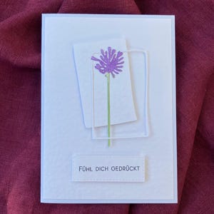 May include: A white greeting card with a textured surface. The card features a purple flower design with a green stem, set within a white rectangular frame. The text "F&Uuml;HL DICH GEDR&Uuml;CKT" is printed at the bottom.
