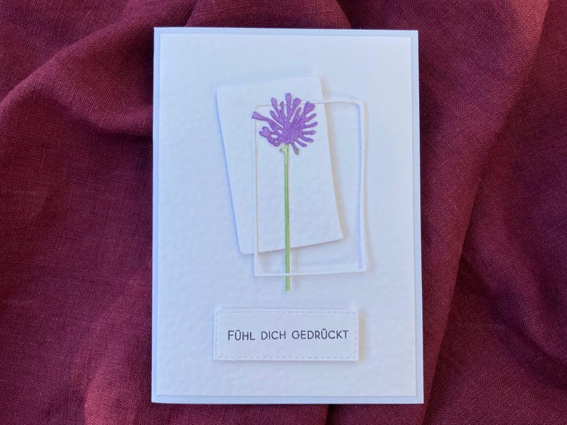 May include: A white greeting card with a textured surface. The card features a purple flower design with a green stem, set within a white rectangular frame. The text "F&Uuml;HL DICH GEDR&Uuml;CKT" is printed at the bottom.