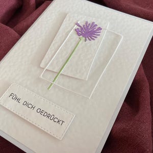 May include: A handmade greeting card with a textured white surface. It features a purple flower with a green stem, set against clear, rectangular frames. The card includes the text "F&Uuml;HL DICH GEDR&Uuml;CKT" on a white label.