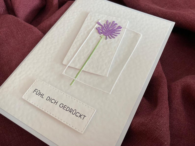 May include: A handmade greeting card with a textured white surface. It features a purple flower with a green stem, set against clear, rectangular frames. The card includes the text "F&Uuml;HL DICH GEDR&Uuml;CKT" on a white label.