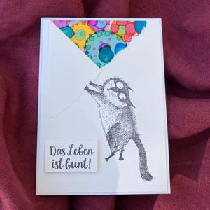 May include: A white greeting card with a whimsical illustration of a cat holding onto a colorful balloon. The text "Das Leben ist bunt!" is printed on a small white rectangle. The background is a deep red fabric.