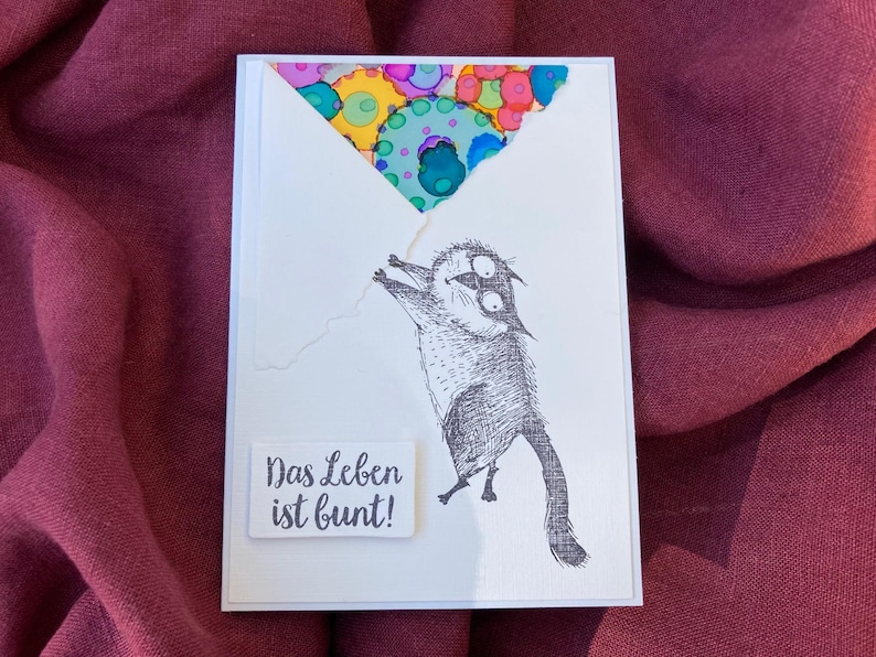 May include: A white greeting card with a whimsical illustration of a cat holding onto a colorful balloon. The text "Das Leben ist bunt!" is printed on a small white rectangle. The background is a deep red fabric.