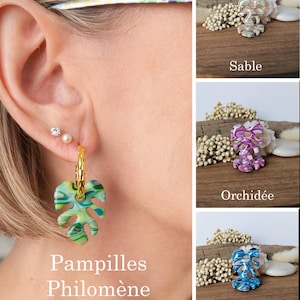 Pode incluir: Brincos de argola dourados com um pingente em forma de folha verde e azul. Os brincos fazem parte da coleção "Pampilles Philomène", com o texto "Jardin tropical" abaixo. Brincos adicionais na imagem são rotulados como "Sable", "Orchidée" e "Lagon".