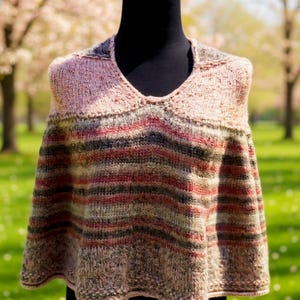 May include: A knitted poncho in shades of pink, red, green, and brown, displayed on a black mannequin. The poncho features a V-neck and horizontal stripes, creating a cozy and stylish look. The background shows a green lawn and trees.