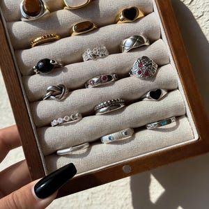 Puede incluir: Una colección de anillos variados expuestos en una caja de joyería de madera. Los anillos presentan diversos diseños, incluyendo engastes de piedras preciosas, bandas texturizadas y detalles en forma de corazón. La paleta de colores incluye tonos plateados, dorados, negros y marrones.