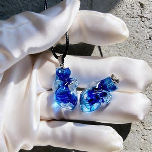 Könnte beinhalten: Zwei herzförmige Anhänger. Einer hängt an einer schwarzen Kordelkette. Die Anhänger sind transparent mit leuchtend blauen Wirbeln. Die Anhänger haben silberfarbene Metallbeschläge.