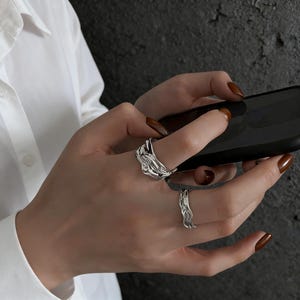 Op de afbeelding: Twee zilveren ringen met een organisch, vloeiend ontwerp worden op een hand getoond. De ringen hebben een textuur van gesmolten metaal. De persoon houdt een zwarte smartphone vast. De achtergrond is een donker, getextureerd oppervlak.
