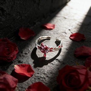 Op de afbeelding: Een verstelbare zilverkleurige ring met een rood rozenontwerp. De ring heeft een centrale roos met een hangende rode edelsteen en rode emaille accenten. Rode rozenblaadjes zijn verspreid rond de ring op een donkergrijze ondergrond.