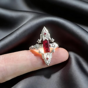 May include: A silver ring with a gothic cross design, featuring a large rectangular red gemstone at the center and smaller red gems. The ring has star and ornate detailing.