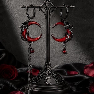 Op de afbeelding: Een paar zwarte oorringen met rode maansikkelhangers en kleine rode druppelvormige hangers. Eén oorbel heeft een lange, dunne zwarte ketting. De oorbellen hangen aan een zwarte metalen standaard met een motief van een maansikkel en rozen.