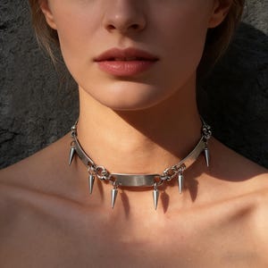 May include: A silver choker necklace with a flat bar design and dangling cone-shaped spikes. The necklace is made of a shiny metal and has a chain-link style. The spikes are evenly spaced along the bottom of the bar.