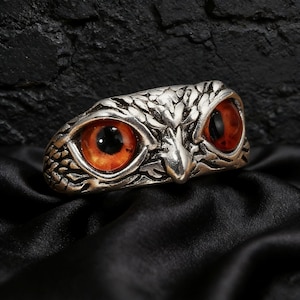 May include: A silver owl ring with intricate feather detailing. The owl's eyes are a striking orange with black pupils. The ring is designed to be worn on a finger, displaying the owl's face.