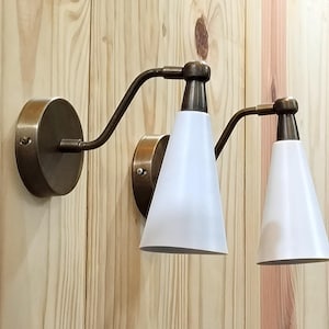 May include: A pair of wall-mounted sconces with a vintage aesthetic. Each sconce features a circular, bronze-colored base, a curved arm, and a white, cone-shaped shade. The fixtures are mounted on a light-colored wooden wall.