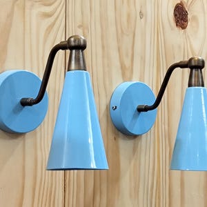 Könnte beinhalten: Zwei hellblaue Wandleuchten mit konischem Schirm und einem dunkelbronzenen Arm und Leuchte. Die Leuchten sind auf einer hellen Holzoberfläche montiert. Die Leuchten haben ein modernes Design aus der Mitte des Jahrhunderts.