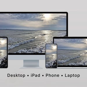 Op de afbeelding: Een desktopcomputer, iPad, telefoon en laptop tonen een schilderachtig uitzicht op de oceaan onder een bewolkte hemel. De afbeelding toont golven die op een rotsachtige kustlijn breken, met de zon die door de wolken schijnt. De apparaten staan op een lichtgrijze ondergrond.